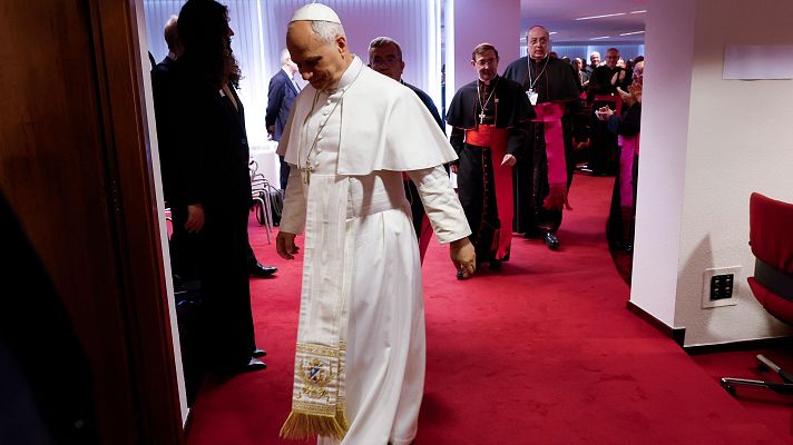 El Papa Francisco avanza por un pasillo con alfombra roja, acompañado por clérigos. Una mujer se asoma a la izquierda.
