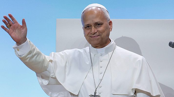 Primer plano del Cardenal Angelo Comastri, sonriendo y saludando con la mano derecha. Viste sotana blanca, solideo y crucifijo pectoral. Fondo dividido en azul y blanco/gris claro. Micrófono visible.