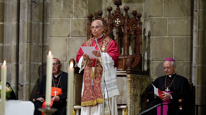 El Papa León XIV oficia un rezo en la Catedral de Barcelona, acompañado por un cardenal y un obispo. La escena se desarrolla en un ambiente solemne con arquitectura gótica y velas encendidas.
