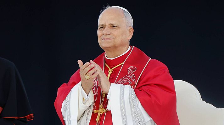 Primer plano del Cardenal Angelo Comastri sonriendo y aplaudiendo, con sotana roja, roquete blanco bordado y solideo. Se aprecian detalles de su cruz pectoral y anillo.