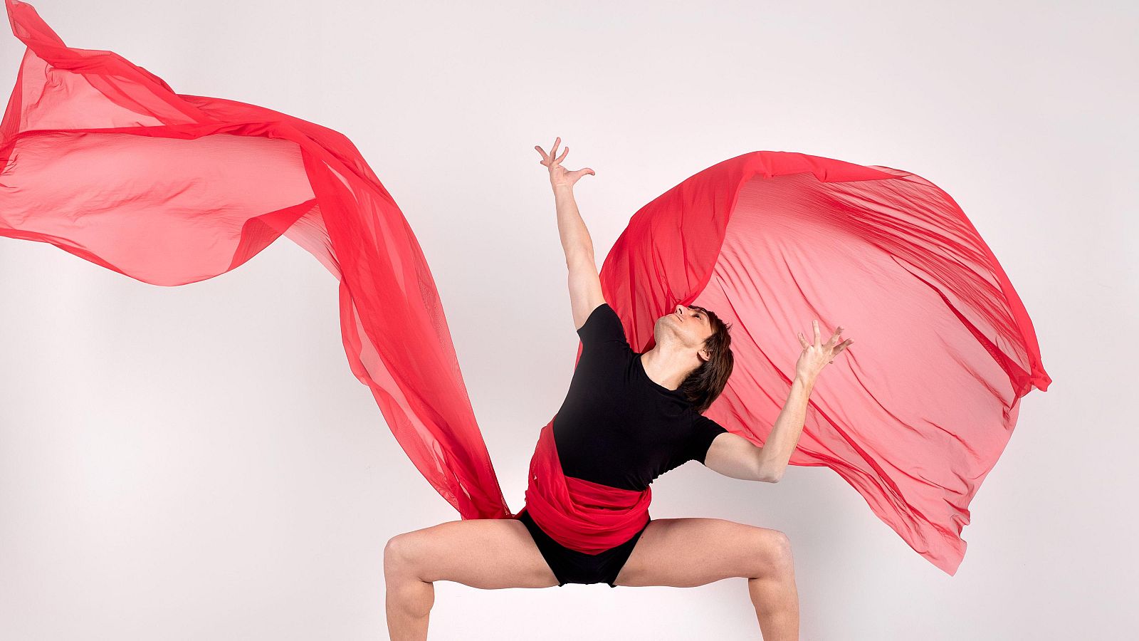 Gonzalo, en pose de danza, luce camiseta y bañador negros, con una tela roja translúcida ondeando desde su cintura como alas sobre fondo claro.