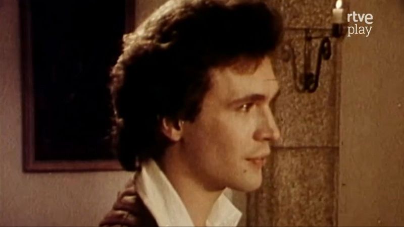 Miguel Bos� de perfil, con cabello oscuro y rizado, camisa blanca y prenda exterior oscura. Fondo con vela encendida y logo de rtve play. Escena de "El juglar y la reina".