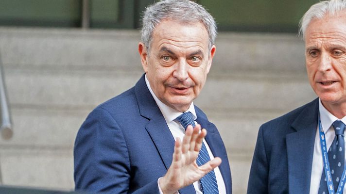 José Luis Rodríguez Zapatero, expresidente del Gobierno de España, en primer plano con traje azul y corbata, levanta la mano derecha. A su derecha, otro hombre con traje y acreditación.