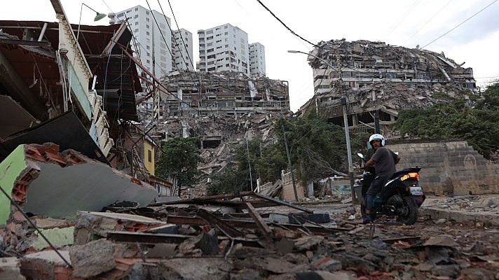 Ruinas de edificios y escombros cubren el suelo, con una persona en motocicleta en medio de la destrucción. Edificios modernos intactos al fondo.
