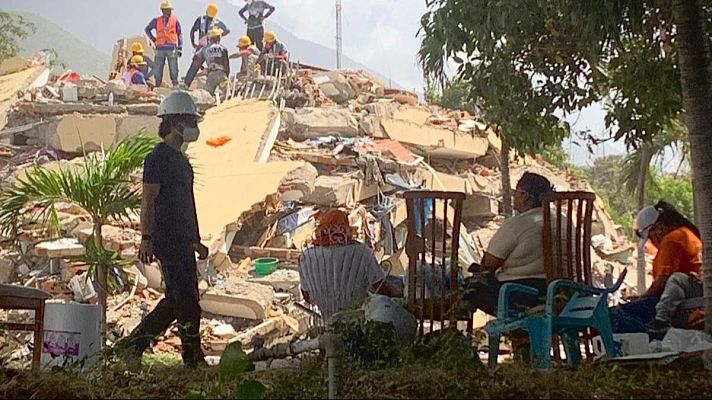 Equipo de rescate en acción sobre escombros de edificio colapsado. Personal con cascos y chalecos naranjas, y observadores en primer plano. Entorno exterior con vegetación.