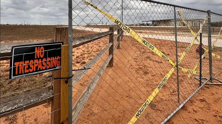 Paisaje exterior con vallas de tela metálica y madera, señalizadas con \"CAUTION\" y \"NO TRESPASSING\" sobre un suelo de tierra rojiza bajo un cielo nublado.