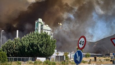 Helic�ptero amarillo con balde de agua rojo combate incendio forestal. Humo negro domina el cielo sobre monta�as. Edificio industrial verde con "VIA" a la izquierda. Se�ales de tr�fico visibles.