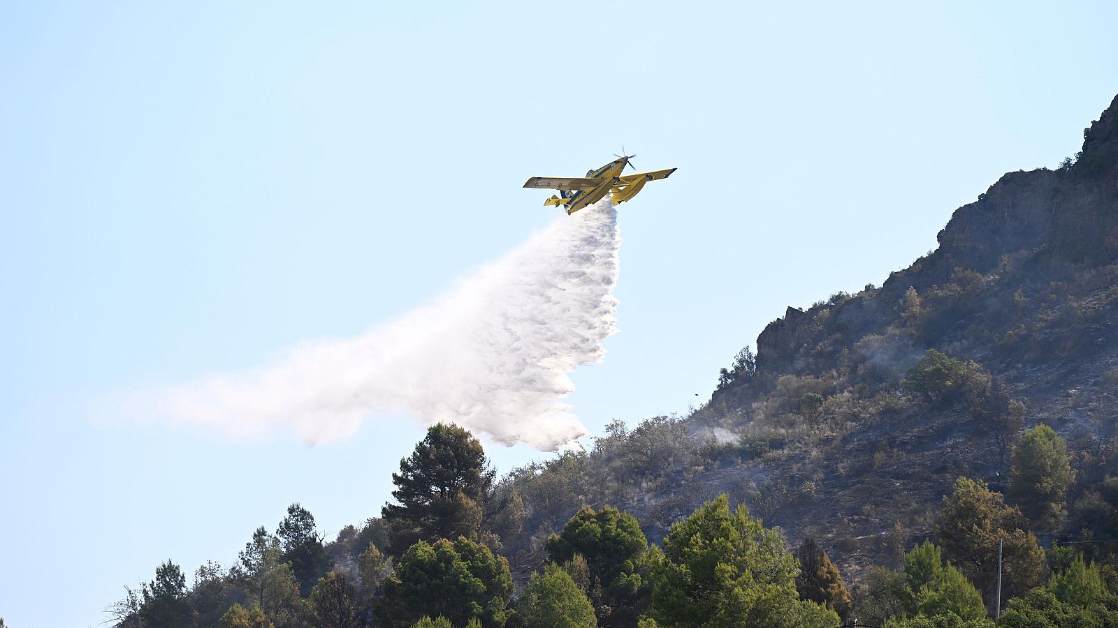 Avión anfibio amarillo descarga agua sobre incendio forestal en ladera montañosa. Vegetación quemada y verde se alternan bajo cielo azul.