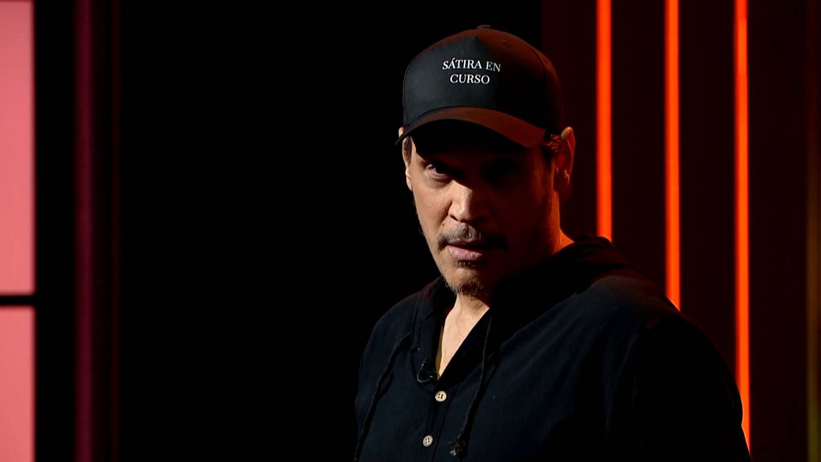 Roberto Peris Mencheta, actor español, con gorra "SÁTIRA EN CURSO" en un escenario con luces rojas anaranjadas. Expresión seria y concentrada.