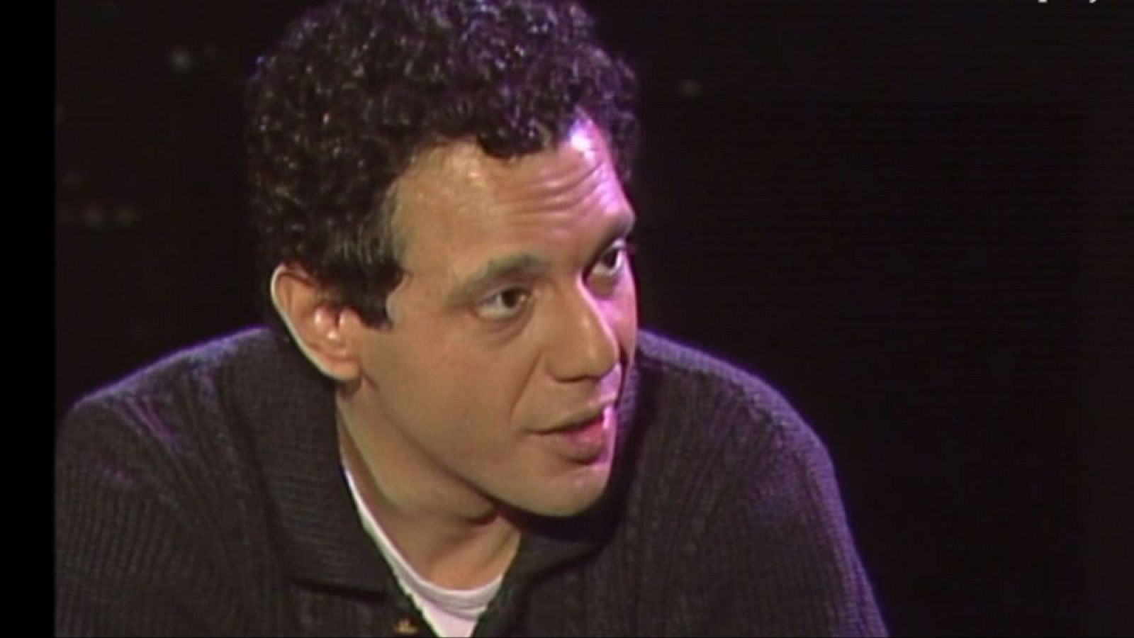 Lluís Pasqual, director de teatro, de perfil, con expresión concentrada y boca entreabierta. Cabello oscuro y rizado, viste prenda oscura sobre blanco. Fondo desenfocado con luz púrpura.