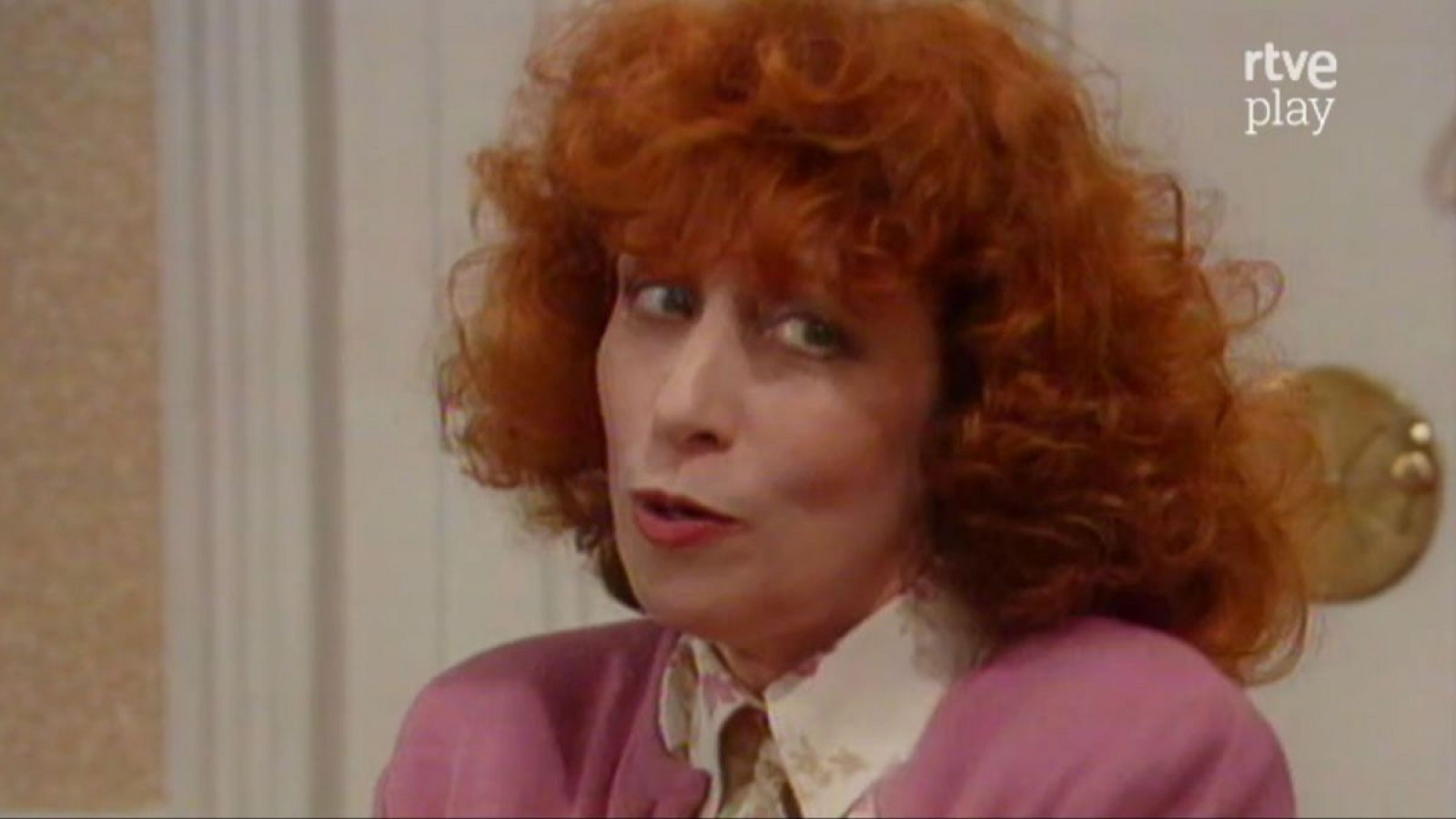 Amparo Baró, actriz española, en primer plano con cabello pelirrojo y rizado, ojos verdes y boca entreabierta. Viste chaqueta rosa y camisa blanca. Fondo desenfocado con logo 'rtve play'.