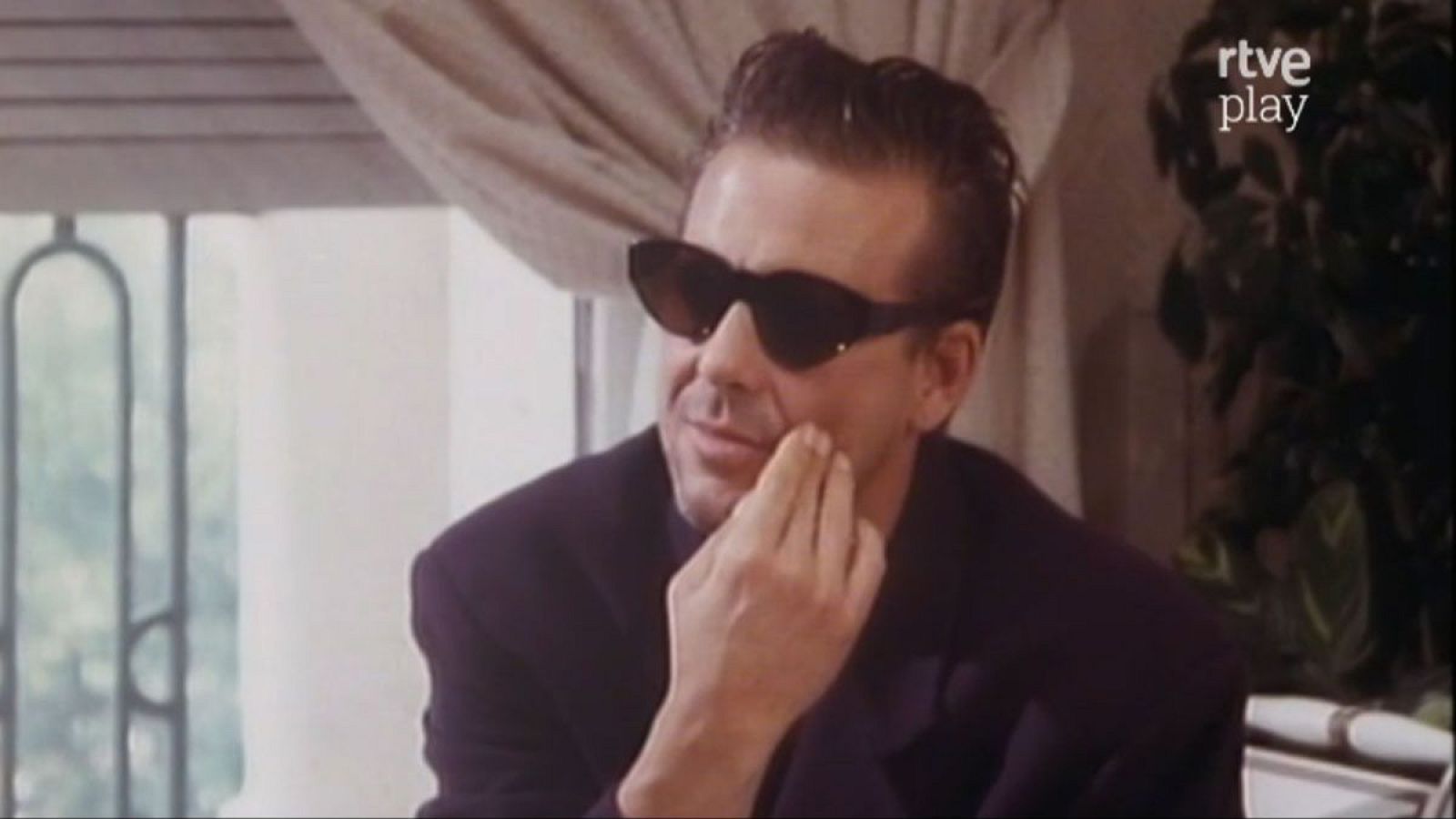 Mickey Rourke, actor, con gafas oscuras y chaqueta, posa pensativo. Fondo con ventana, planta y logo 'rtve play'.
