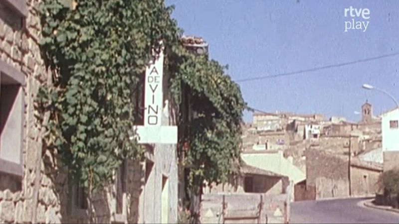 Edificio con cartel "DE VINO" y vegetaci�n, pueblo en colina con torre de iglesia al fondo. Logo "rtve play" en esquina superior derecha.