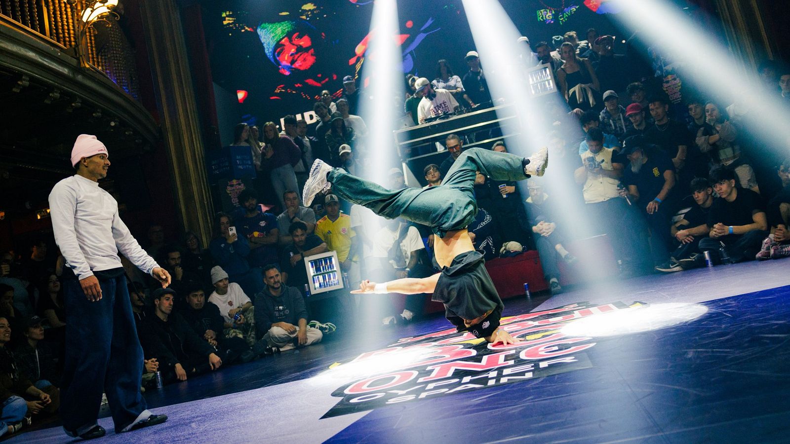 Bailarín de breakdance en acción en el "RED BULL BC ONE CYPHER SPAIN", con público y equipo de DJ en un recinto con balcones de madera oscura y cortinas rojas.