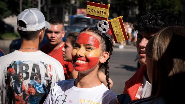 Joven con rostro pintado de rojo y amarillo, diadema con banderas de España y balón de fútbol, sonríe. Fondo festivo con aficionados al deporte y símbolos de España y la NBA.