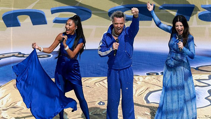 Nicole Scherzinger, Robbie Williams y otra mujer cantan en un escenario con fondo de pantalla de \"26\" y paisaje marino. Todos visten de azul y sostienen micrófonos.