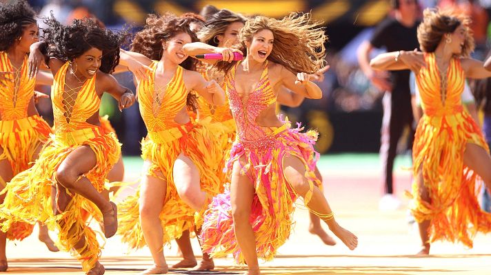 Shakira canta con un micrófono rosa, luciendo un traje de flecos rosa, amarillo y naranja, rodeada de bailarinas en un evento al aire libre.