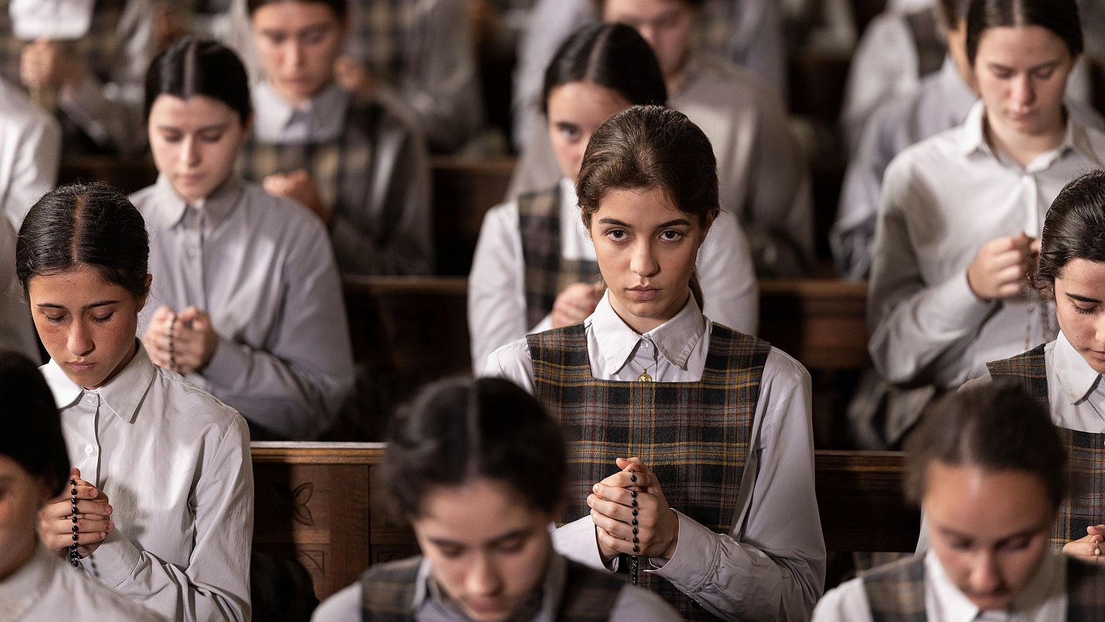 Jóvenes mujeres con uniformes a cuadros y camisas blancas, sentadas en filas, algunas rezando con rosarios en un entorno solemne.