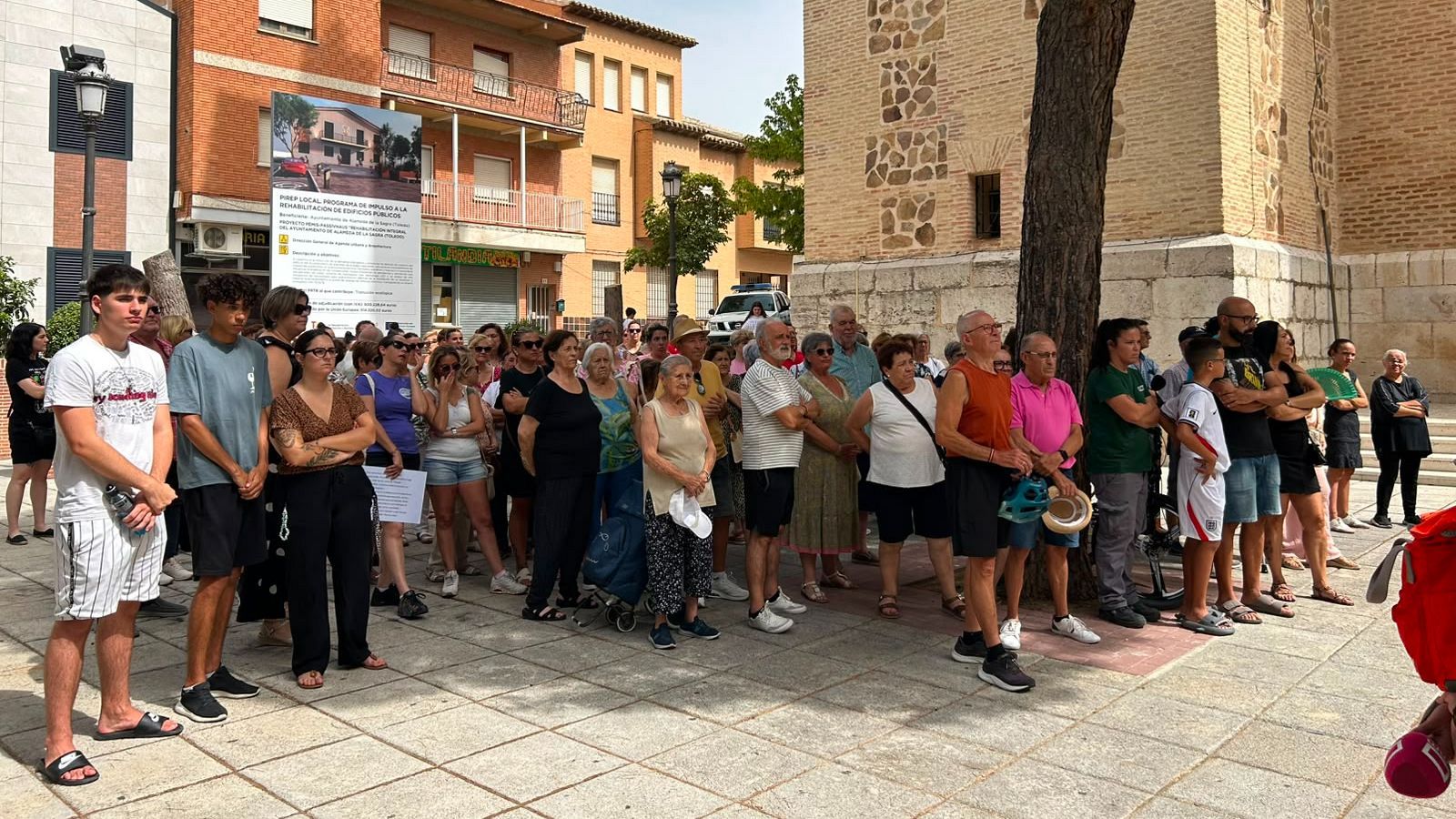 Multitud solemne en plaza soleada, con cartel de "PLAN LOCAL PROYECTO DE MEJORA DE LA REHABILITACIÓN DE ESPACIOS PÚBLICOS".