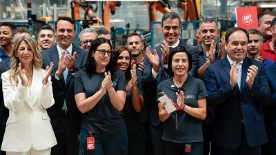 Pedro S�nchez, Yolanda D�az y Jim Baumbick celebran en una planta de Ford rodeados de maquinaria industrial y otros asistentes.