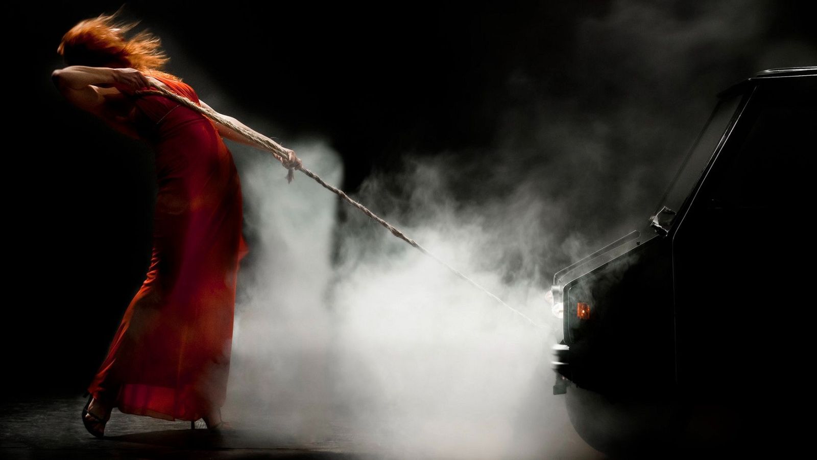 Mujer de cabello rojizo y vestido rojo tira de una cuerda hacia un vehículo oscuro envuelto en humo blanco, bajo una luz dramática. "Laboratorio escénico".