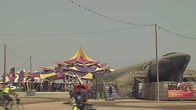 Festival al aire libre con carpa colorida, estructura "OCB MONEGROS" tipo submarino, personas en bicicleta y cartel de Repsol en entorno des�rtico.