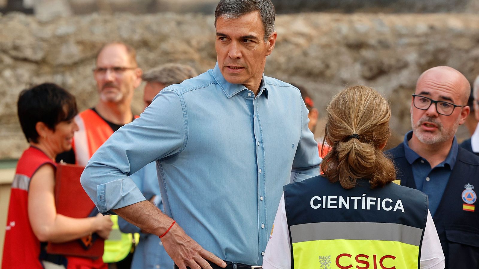 Pedro Sánchez, con camisa vaquera y pulsera roja, es observado por personal del CSIC y Protección Civil en un terreno irregular.