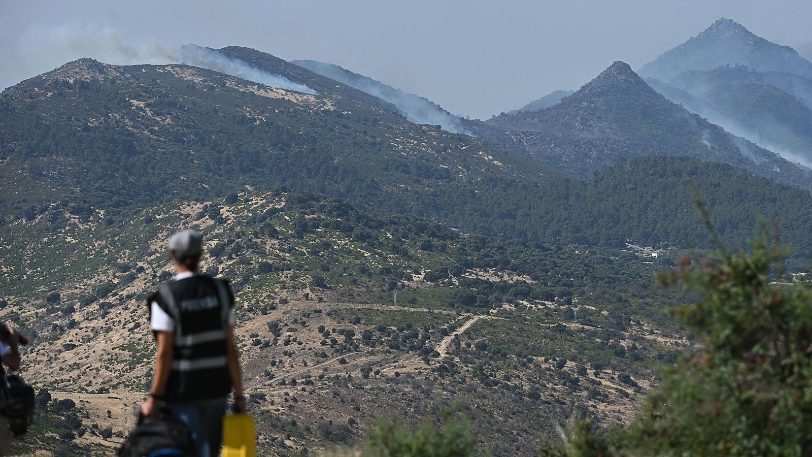 Persona con chaleco de "PRENSA" observa un paisaje montañoso con columnas de humo, posiblemente cubriendo un incendio forestal.