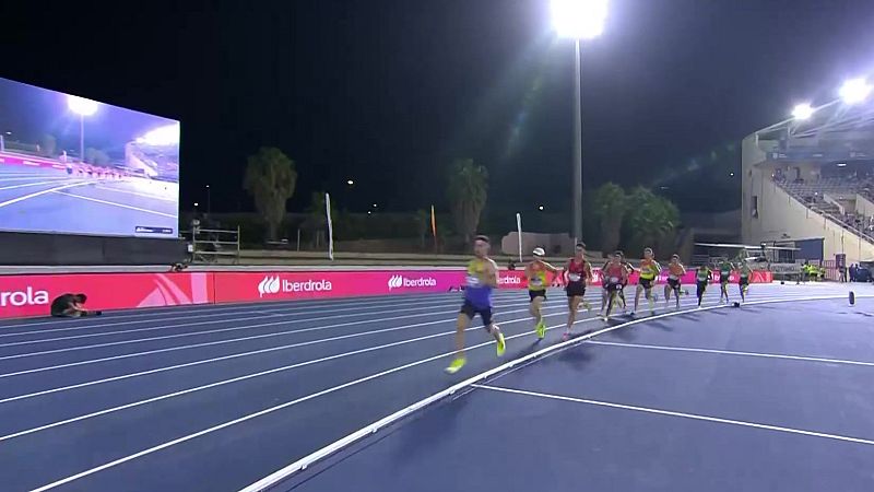 Atletas masculinos compiten en una carrera de atletismo nocturna en una pista azul, con vallas de Iberdrola y una pantalla LED visible.