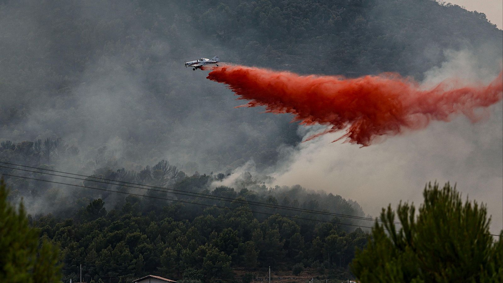 Avión de extinción de incendios lanza líquido rojo sobre un bosque en llamas, cubierto de humo gris. Líneas eléctricas y tejado de edificio visibles.
