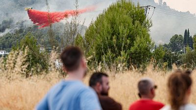 Avi�n amarillo descarga retardante rojo sobre incendio forestal con casas y monta�as. Cuatro personas observan desde un campo de hierba seca.