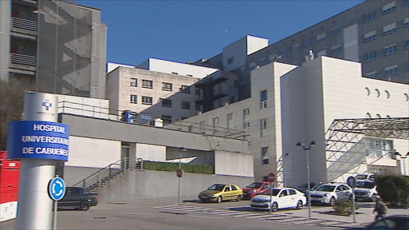 Vista exterior del Hospital Universitario de Cabueñes con vehículos y un cartel anunciador en primer plano bajo un cielo azul.