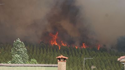 Incendio forestal con llamas intensas y humo denso sobre un bosque, amenazando estructuras habitadas en primer plano.