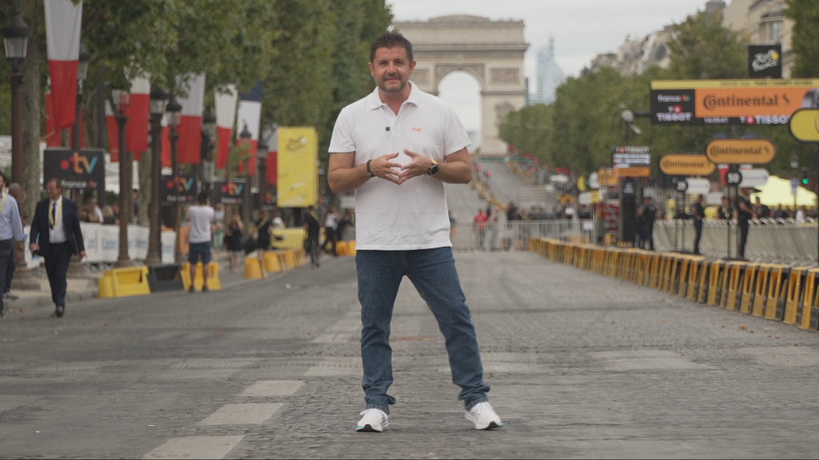 Carlos de Andrés, periodista de RTVE/Teledeporte, posa en los Campos Elíseos de París, con el Arco del Triunfo de fondo. Lleva polo blanco, vaqueros y zapatillas, y mira al frente. La avenida está engalanada con banderas francesas y carteles de patrocinadores.