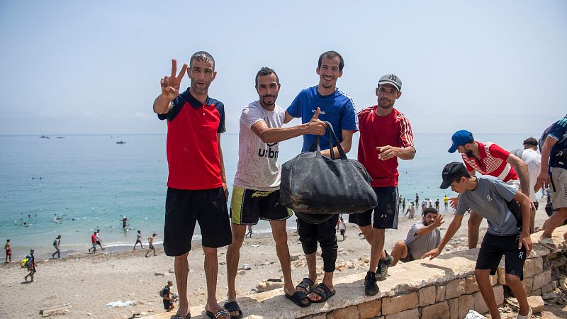 Hombres sonríen y celebran en una playa concurrida, con el mar turquesa y el cielo azul de fondo. Uno levanta el pulgar, otro hace el signo de la victoria.