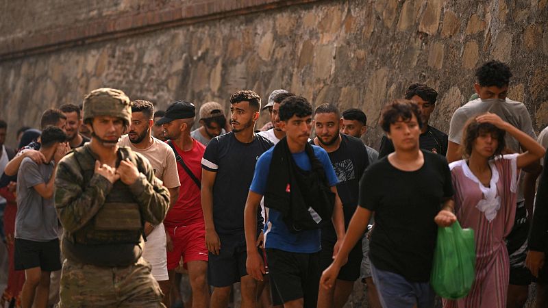 Un grupo diverso de personas, incluyendo un militar, caminan a lo largo de un muro de piedra en una escena diurna. La multitud se desplaza junta, con variadas vestimentas y expresiones.
