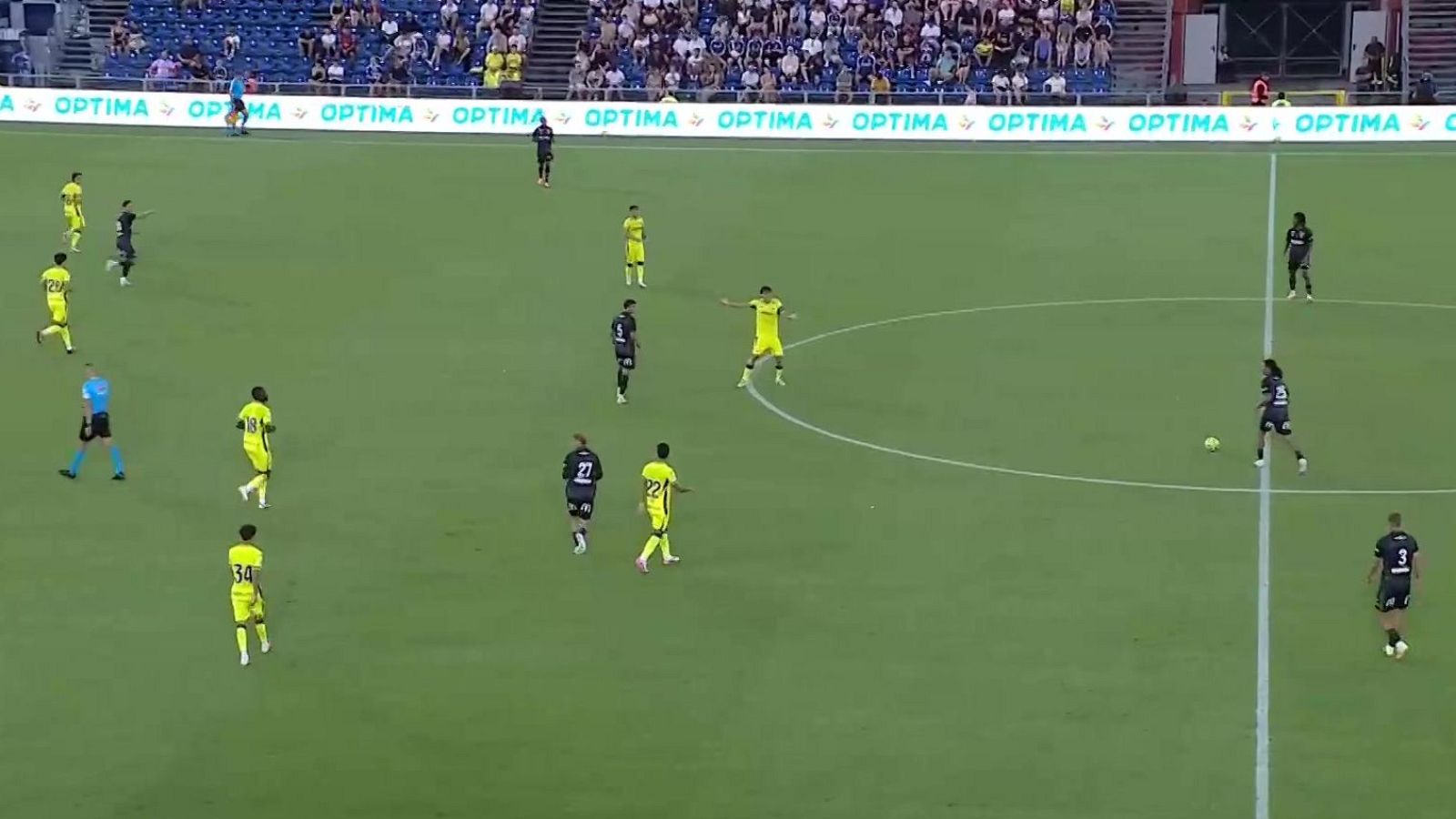 Vista aérea de un partido de fútbol con jugadores de uniformes amarillos/verdes y oscuros, un árbitro azul, césped verde, gradas y valla publicitaria "OPTIMA".