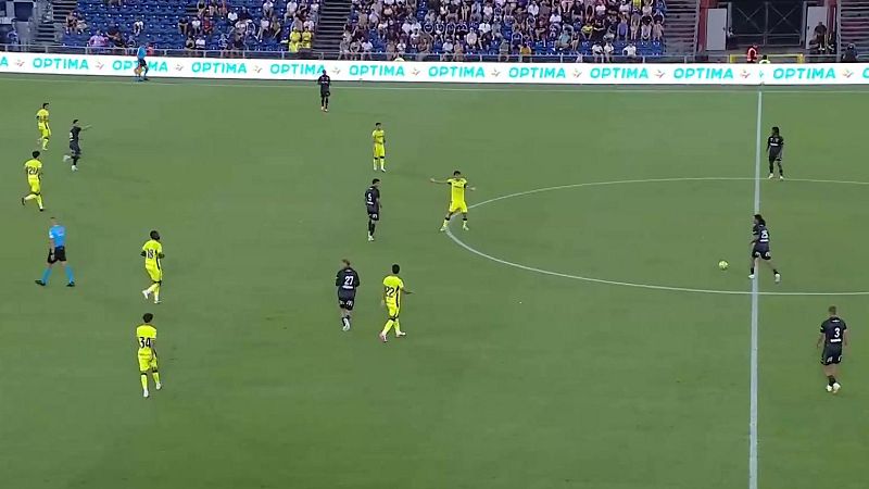 Vista a�rea de un partido de f�tbol con jugadores de uniformes amarillos/verdes y oscuros, un �rbitro azul, c�sped verde, gradas y valla publicitaria "OPTIMA".