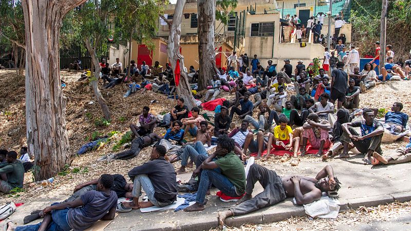Gran grupo de hombres jóvenes y adultos, principalmente de ascendencia africana, descansan en un área exterior con suelo de tierra y grava, rodeados de árboles y edificios modestos al fondo.