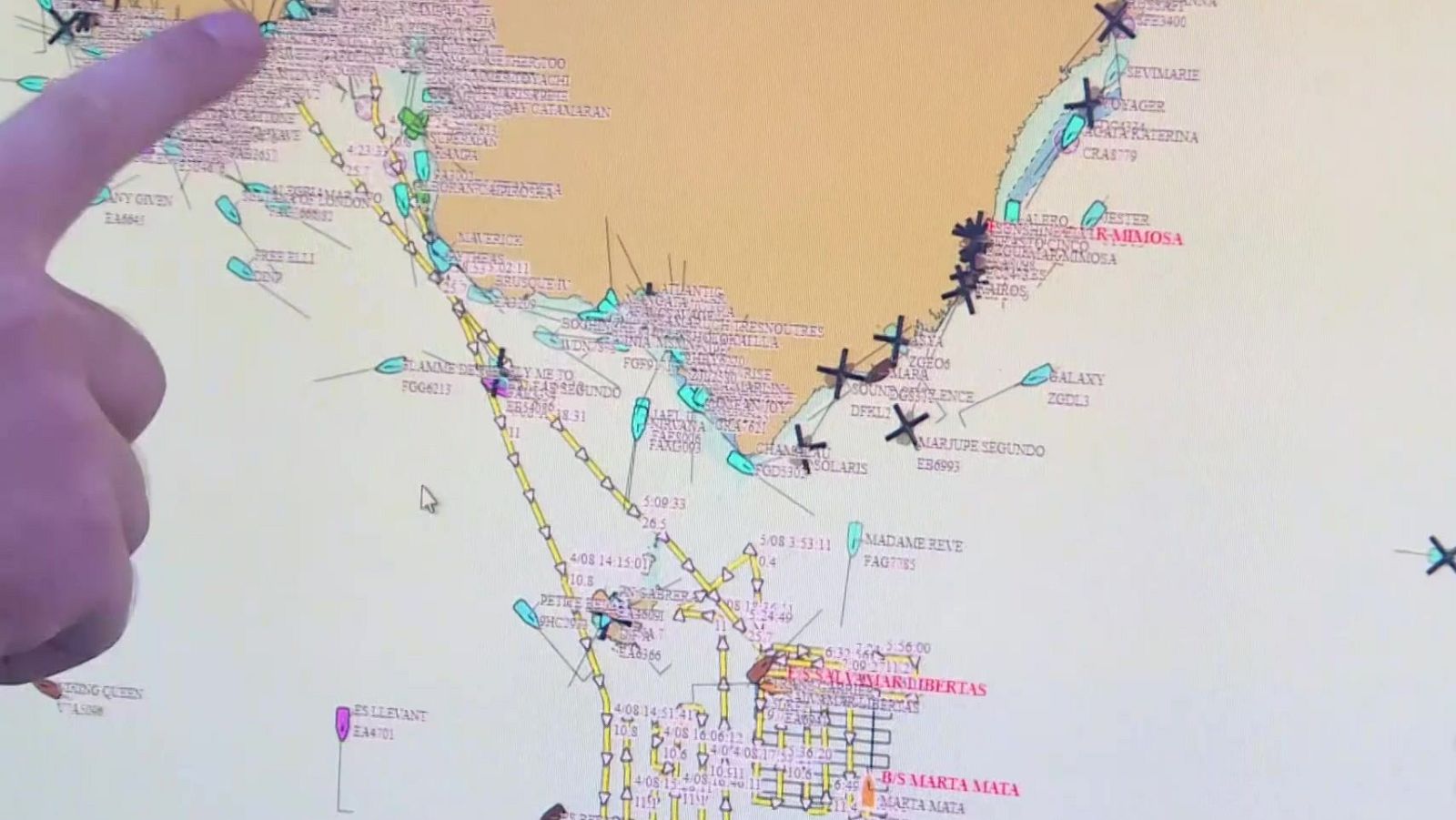 Pantalla digital con mapa náutico, embarcaciones, rutas y marcas de tiempo. Dedo señalando el mapa. Nombres de embarcaciones visibles: ANY COVEN, ZAMU, VOYAGER, RAIMOSA (rojo).