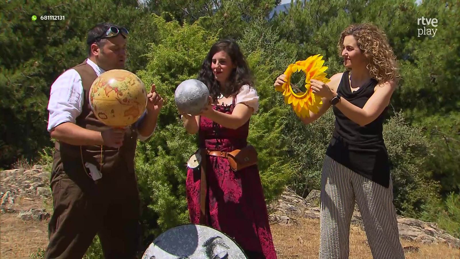 Un hombre, una mujer y otra mujer en un entorno natural, sosteniendo un globo terráqueo, un modelo lunar y un accesorio de girasol/sol, respectivamente. Se observa el logo 'rtve play' y un número de teléfono.