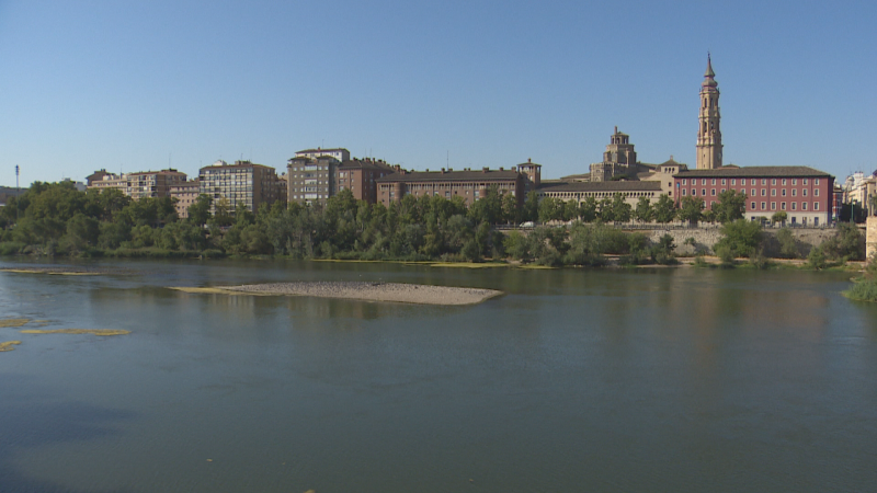 Vista panorámica de Zaragoza desde el río Ebro, con la Basílica del Pilar y edificios modernos en el horizonte bajo un cielo azul despejado.