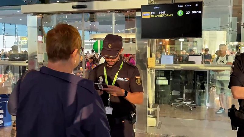 Agente de Policía Nacional revisa un documento a una persona en un aeropuerto. Pantalla muestra vuelo FR 3142 a MILAN/BERGAMO y cinta 23 a las 08:18.