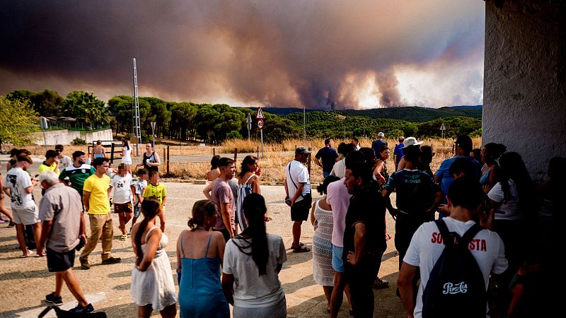Multitud observa incendio forestal con densa columna de humo. "SOSTA" y "ALONSO" visibles en camisetas. Paisaje con colinas, terreno árido y torre de comunicaciones.