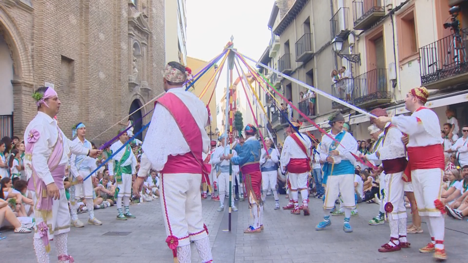 Danza de cintas folclórica en calle histórica con hombres vestidos de blanco y fajas de colores, tejiendo cintas de múltiples tonos alrededor de un poste central. Espectadores en balcones y a pie de calle.