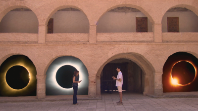 En un patio con arcos de ladrillo, una mujer y un hombre observan eclipses solares representados en los arcos, interactuando con sus dispositivos m�viles.