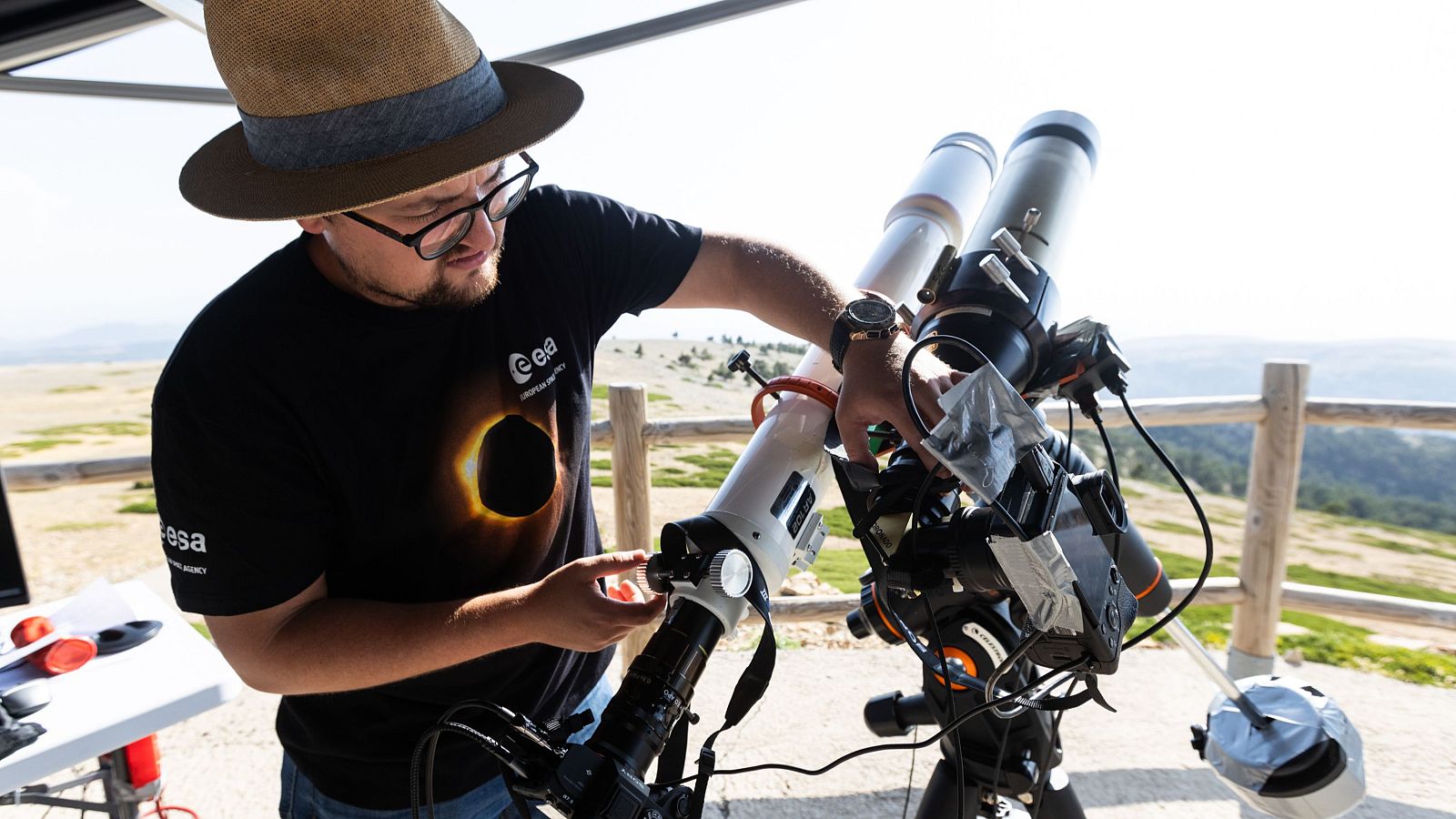 Hombre con camiseta de la ESA y sombrero de paja ajusta telescopio para observar un eclipse solar total en un paisaje natural.
