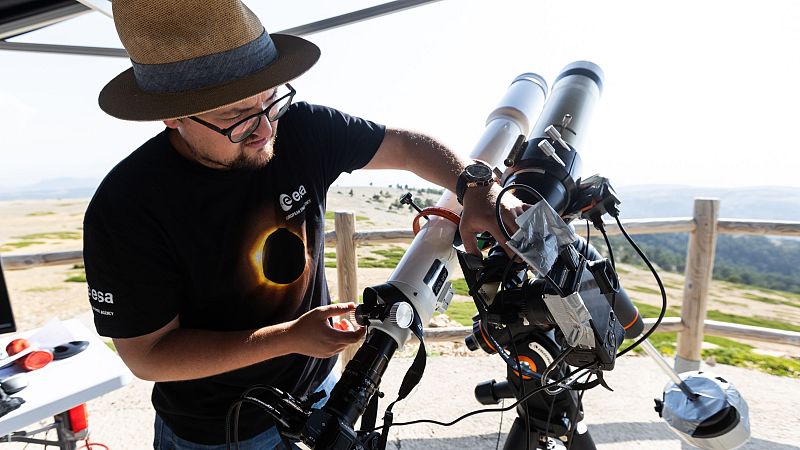 Hombre con camiseta de la ESA y sombrero de paja ajusta telescopio para observar un eclipse solar total en un paisaje natural.