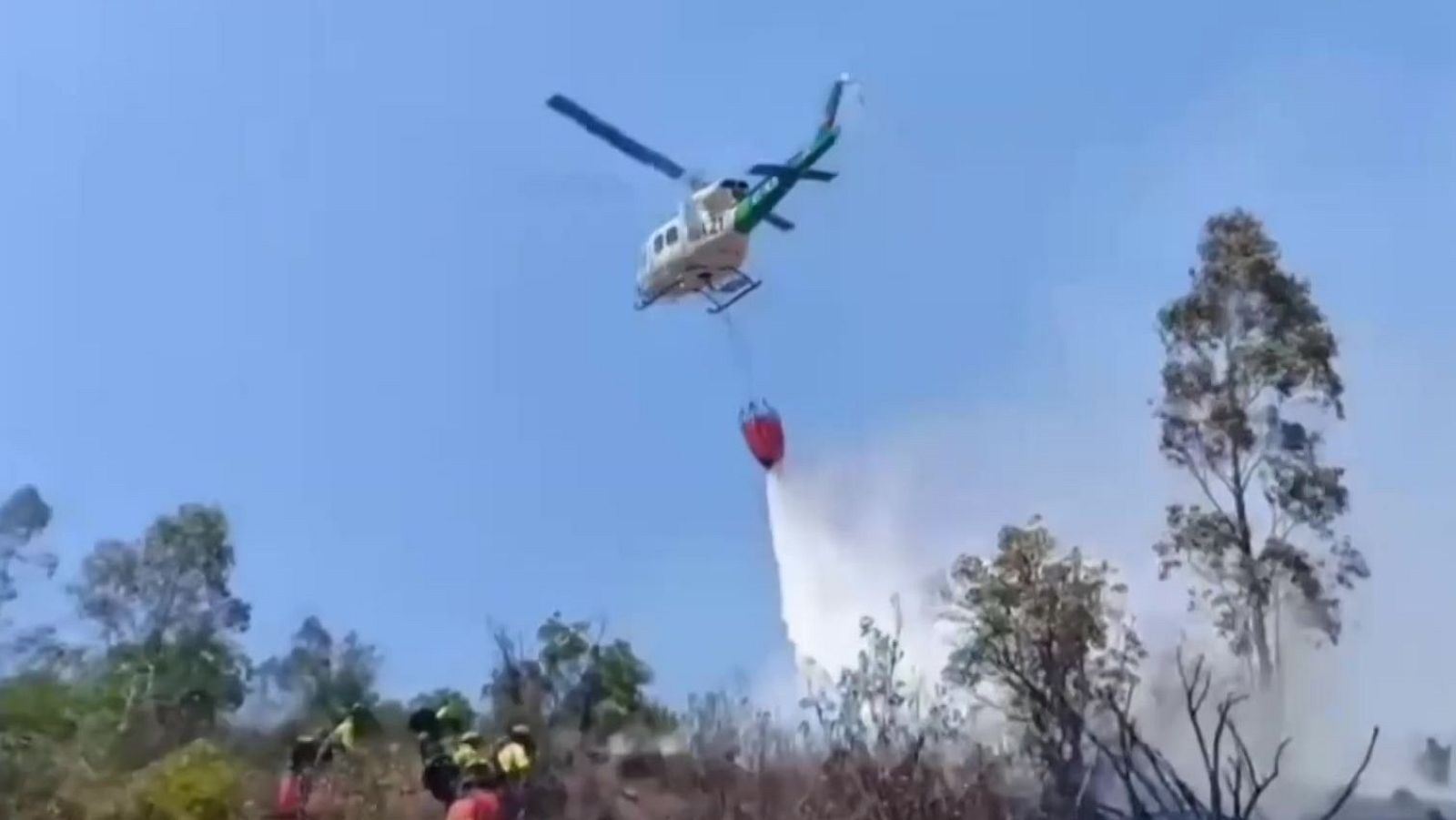 Helicóptero descarga agua sobre incendio forestal. Bomberos en acción en terreno quemado. Humo blanco visible.