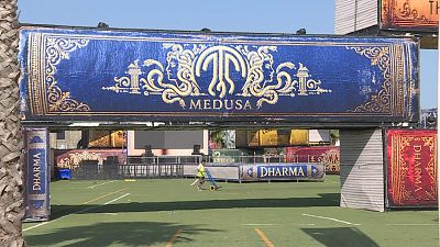 Arco de entrada tem�tico de libros gigantes con "MEDUSA" y "DHARMA" en dorado. Persona con camiseta amarilla sobre c�sped artificial.